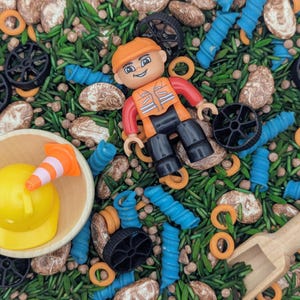 May include: A construction-themed sensory bin with a yellow hard hat, orange and white traffic cone, and a construction worker figurine. The bin also contains black wheels, blue pasta shapes, and wooden scoop.