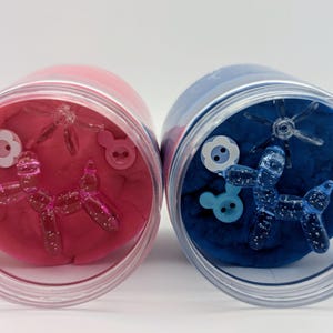 May include: Two clear plastic jars filled with pink and blue playdough. Each jar contains decorative elements: a balloon dog, flower and button-shaped embellishments. The pink jar has pink accents, while the blue jar has blue accents.