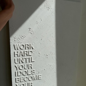 May include: A rectangular white canvas featuring a heavily textured, impasto-style surface. The words WORK HARD UNTIL YOUR IDOLS BECOME YOUR RIVALS are embossed in raised, capital letters on the lower left side of the textured white background.