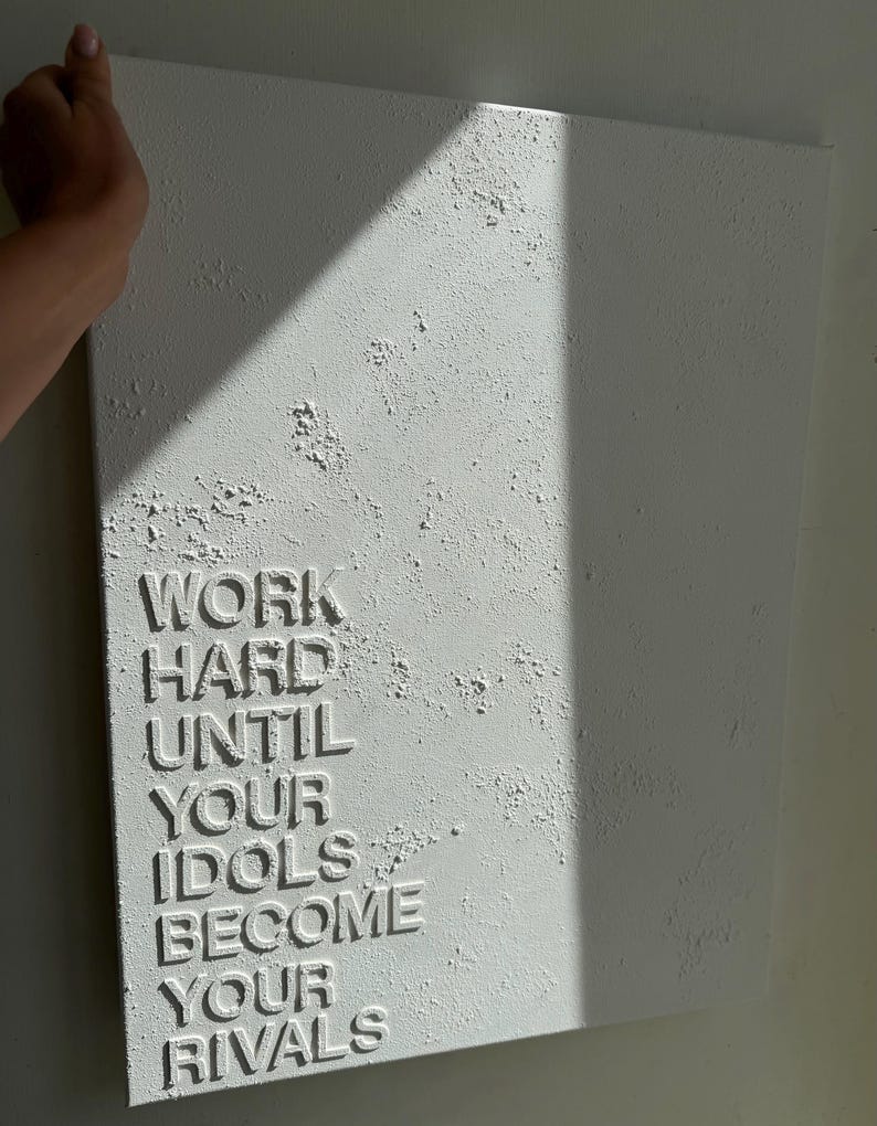 May include: A rectangular white canvas featuring a heavily textured, impasto-style surface. The words WORK HARD UNTIL YOUR IDOLS BECOME YOUR RIVALS are embossed in raised, capital letters on the lower left side of the textured white background.