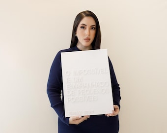 May include: A rectangular white sign featuring embossed text that reads O IMPOSSÍVEL É UM EMARANHADO DE PEQUENOS POSSÍVEIS. The sign is held against a dark navy blue long-sleeved garment, creating a high-contrast visual display of the lettering.