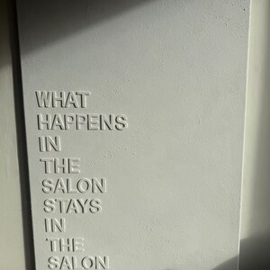 May include: A rectangular white wall art piece featuring raised, embossed lettering that reads: WHAT HAPPENS IN THE SALON STAYS IN THE SALON. The surface has a subtle, textured finish, and the letters cast soft shadows under directional lighting.