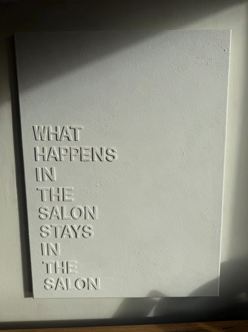 May include: A rectangular white wall art piece featuring raised, embossed lettering that reads: WHAT HAPPENS IN THE SALON STAYS IN THE SALON. The surface has a subtle, textured finish, and the letters cast soft shadows under directional lighting.