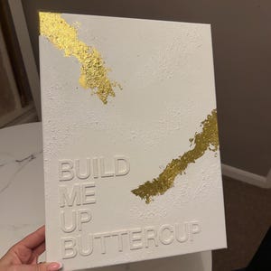 May include: A rectangular white canvas featuring a textured surface with raised white lettering that reads BUILD ME UP BUTTERCUP. The artwork is accented with two diagonal streaks of metallic gold leaf applied to the top left and middle right corners.