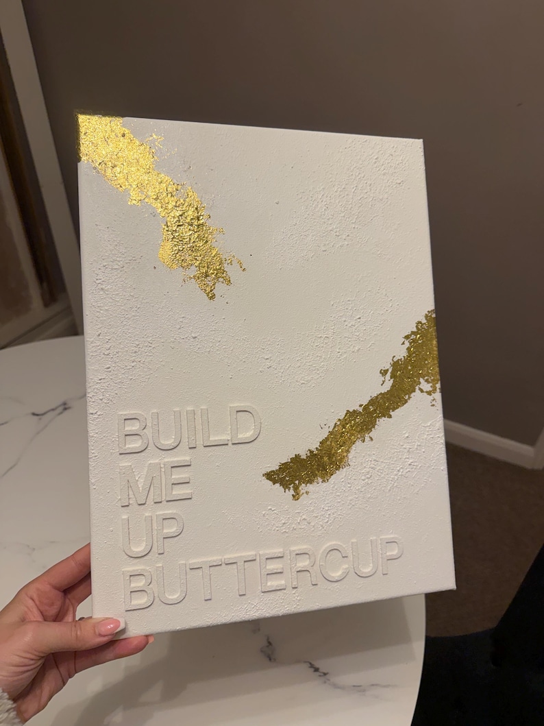 May include: A rectangular white canvas featuring a textured surface with raised white lettering that reads BUILD ME UP BUTTERCUP. The artwork is accented with two diagonal streaks of metallic gold leaf applied to the top left and middle right corners.