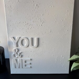 May include: A rectangular white canvas wall art piece featuring a heavily textured, impasto-style surface. The words YOU & ME are displayed in raised, three-dimensional white lettering on the left side of the canvas, creating a monochromatic design.