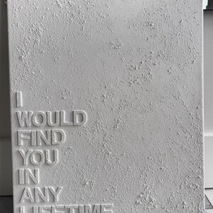 May include: A rectangular white canvas featuring a heavily textured, impasto-style surface. The words I WOULD FIND YOU IN ANY LIFETIME are embossed in a clean, sans-serif font on the left side, contrasting with the rough, raised plaster texture.