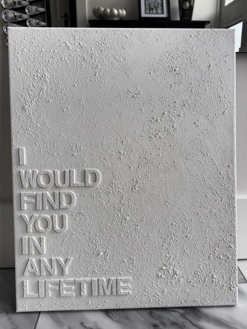 May include: A rectangular white canvas featuring a heavily textured, impasto-style surface. The words I WOULD FIND YOU IN ANY LIFETIME are embossed in a clean, sans-serif font on the left side, contrasting with the rough, raised plaster texture.