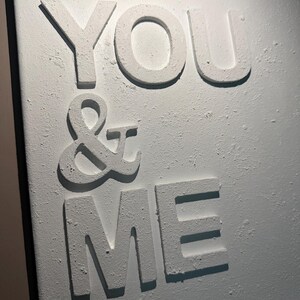 May include: A white wall art piece featuring the raised, textured text "YOU & ME" in capital letters. The surface of the letters and the background has a rough, matte, plaster-like finish, creating a three-dimensional effect with shadows.