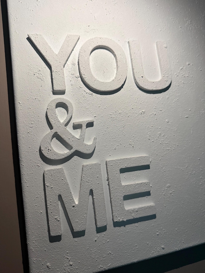 May include: A white wall art piece featuring the raised, textured text "YOU & ME" in capital letters. The surface of the letters and the background has a rough, matte, plaster-like finish, creating a three-dimensional effect with shadows.
