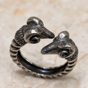 May include: A silver-toned metal ring featuring two ram heads facing each other at the open ends. The band has a textured, ribbed design with intricate spiral detailing near the heads, finished with a dark, oxidised patina in the recessed areas.