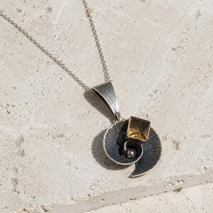 Puede incluir: Un collar con colgante de plata que presenta un diseño en forma de concha en espiral con un acabado oscuro oxidado y líneas finamente grabadas. Una piedra preciosa cuadrada de color ámbar está engastada en el centro junto a una cuenta de plata.