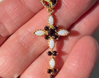 Könnte beinhalten: Eine goldfarbene Halskette mit einem Kreuzanhänger. Der Anhänger hat ein Kreuzdesign mit weißen und roten Edelsteinen. Die Halskette wird von einer Hand gehalten, die die komplizierten Details des Schmucks hervorhebt.