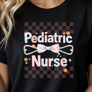 May include: Black t-shirt with the words "Pediatric Nurse" in a colorful design. The design includes a stethoscope, bow, and flowers on a checkered background. The shirt is a crew neck style.