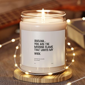 May include: A lit soy candle in a clear glass jar with a white label. The label reads "DARLING... YOU ARE THE MISSING FLAME THAT LIGHTS MY WICK." The candle sits on a round wooden coaster, surrounded by string lights.