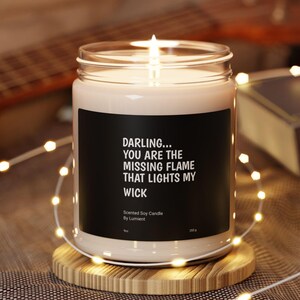 May include: A lit scented soy candle in a glass jar with a black label that reads "DARLING... YOU ARE THE MISSING FLAME THAT LIGHTS MY WICK." The candle sits on a wooden coaster, surrounded by string lights. A guitar and book are in the background.