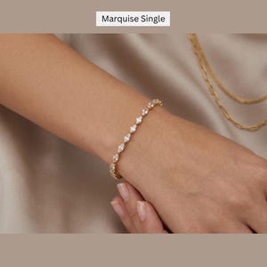 Puede incluir: Una pulsera de tono dorado con una serie de piedras transparentes de corte marquesa dispuestas en línea continua. El texto "Marquise Single" aparece en la parte superior. La pulsera se lleva en la muñeca, con una cadena dorada visible cerca.