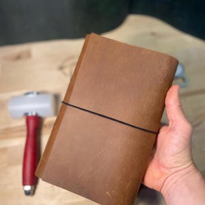 Könnte beinhalten: Ein braunes Lederjournal mit einem schwarzen Gummibandverschluss. Das Journal wird in einer Hand gehalten. Ein rot-weißer Hammer befindet sich im Hintergrund.