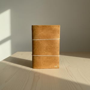 Könnte beinhalten: Ein beigefarbener Notizbuchumschlag aus Leder mit glatter, matter Oberfläche, sichtbarer natürlicher Maserung und Kratzern. Zwei dünne, weiße Gummibänder verlaufen horizontal um den Umschlag. Die Initialen A.B. sind unten rechts eingeprägt.