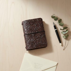 May include: A dark brown leather journal with an intricate embossed floral pattern, secured by a black elastic band. Beside it lies a black fountain pen with a silver tip and a sprig of green eucalyptus leaves, along with a cream-colored envelope.