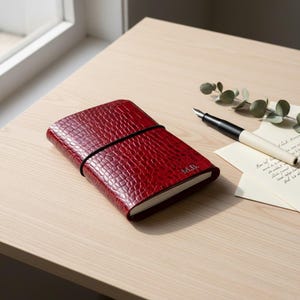May include: A red leather journal with a textured, crocodile-like pattern. The journal is closed with a black elastic band and has the initials "M.B." embossed on the cover. A black and white fountain pen and a sprig of greenery are next to the journal.