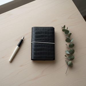 May include: A black notebook with a textured crocodile skin pattern held closed by a thin white elastic band, accompanied by a fountain pen with a cream-coloured barrel and a silver nib, alongside a single sprig of green eucalyptus leaves.