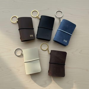 Puede incluir: Cinco pequeños llaveros de cuaderno rectangulares de cuero en marrón oscuro, negro, azul, crema y marrón rojizo. Cada uno cuenta con una anilla de metal, una banda elástica de cierre y algunos tienen iniciales grabadas como FH, FW y NB.