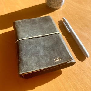 Könnte beinhalten: Ein dunkelgraues Lederjournal mit einem cremefarbenen Gummiband liegt auf einer Holzoberfläche. Das Journal hat die Initialen "S.P." in Gold geprägt. Ein hellgrauer Stift und eine Keramiktasse befinden sich ebenfalls auf dem Tisch.