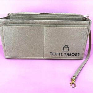 Peut inclure: Organisateur de sac à main en feutre taupe avec plusieurs compartiments et un dessus zippé. L'organisateur a une longue sangle avec un fermoir doré. Le texte "TOTTE THEORY" et une icône de cadenas sont imprimés sur le devant.