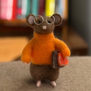 May include: A handcrafted felt mouse figurine wearing an orange sweater and small wire-rimmed glasses. The mouse holds two books, one with a red cover. The mouse has brown fur and pink feet.