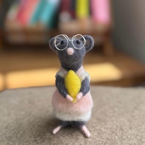Könnte beinhalten: Eine nadelgefilzte graue Mausfigur mit einer drahtgebogenen Brille und einer hellrosa-weißen Weste. Die Maus hält ein leuchtend gelbes, oval geformtes Objekt zwischen ihren Vorderpfoten und hat kleine rosa Füße sowie eine rosa Nase.