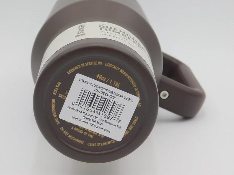 May include: A brown Stanley Quencher tumbler with a handle. The label indicates a 1.18L capacity. The tumbler is designed in Seattle, WA, and ethically manufactured in China. The label also includes a barcode and the text "BPA Free Dishwasher Safe".