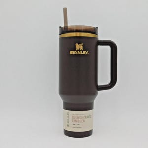 May include: A dark brown Stanley Quencher H2.0 tumbler with a handle and straw. The tumbler has a gold rim and the Stanley logo. The label reads "QUENCHER H2O TUMBLER".
