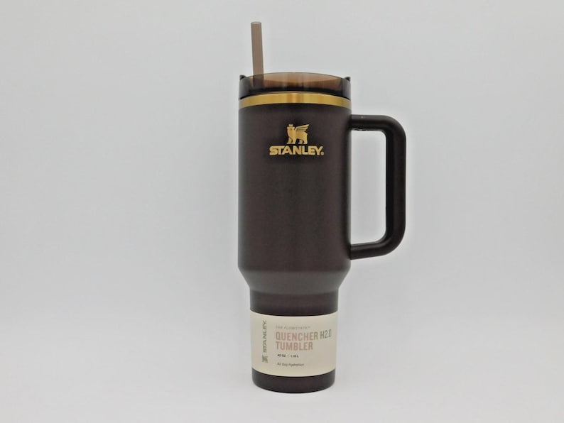May include: A dark brown Stanley Quencher H2.0 tumbler with a handle and straw. The tumbler has a gold rim and the Stanley logo. The label reads "QUENCHER H2O TUMBLER".