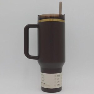 May include: A dark brown insulated travel mug with a handle, a lid, and a straw. The mug has a gold-coloured band near the top and a label near the bottom. The straw is a light brown colour.