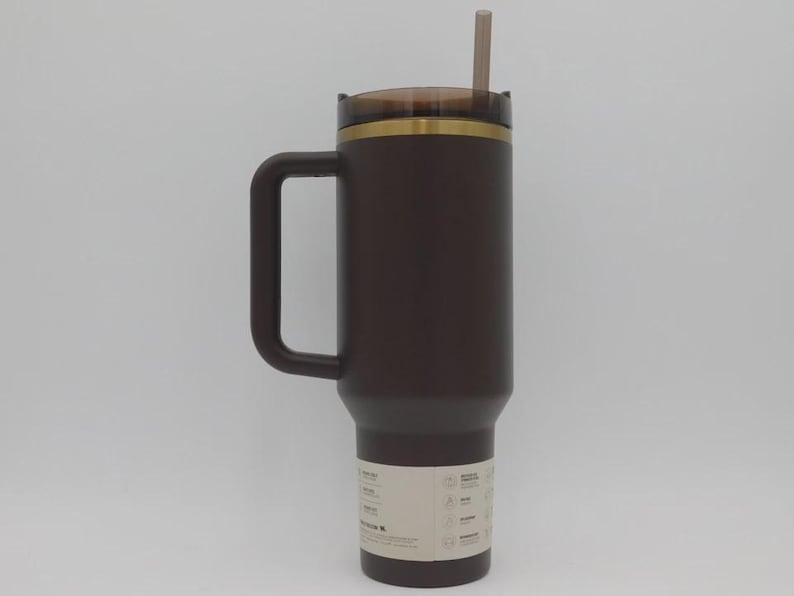May include: A dark brown insulated travel mug with a handle, a lid, and a straw. The mug has a gold-coloured band near the top and a label near the bottom. The straw is a light brown colour.