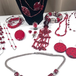 May include: A collection of red jewelry including a multi-strand necklace on a black display, a beaded tassel pendant, a silver-tone chain necklace with red rectangular stones, red beaded bracelets, circular fabric earrings, and a small rose pin.