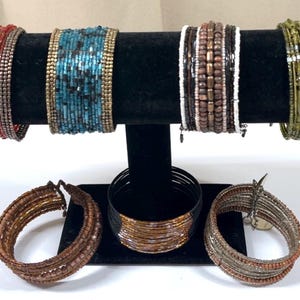 May include: An assortment of beaded bracelets in various colors, including red, turquoise, brown, and green, displayed on a black velvet stand. Several bracelets are also arranged at the base of the stand, showcasing different designs and materials.