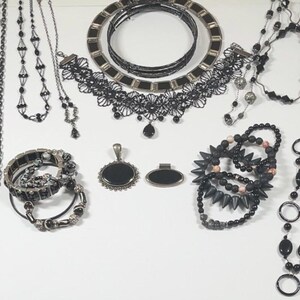 May include: A collection of black necklaces, bracelets, and pendants. The necklaces vary in style, including beaded, chain, and lace designs. Bracelets include studded and beaded styles. Pendants are oval and rectangular.