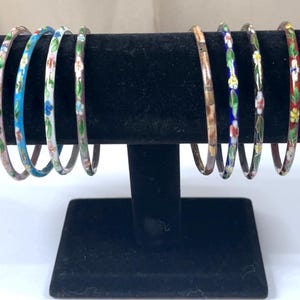 May include: A collection of eight thin, decorative bangles with floral designs. The bangles are displayed on a black stand. The bracelets feature a variety of colors, including pink, blue, white, green, and red, with intricate floral patterns.