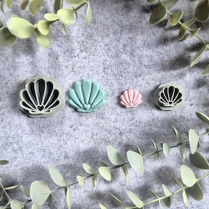 Op de afbeelding: Vier schelpvormige koekjessnijders in verschillende maten en kleuren: zwart, teal en roze. De uitstekers zijn gerangschikt op een grijze ondergrond, omringd door groene bladsprieten. Ideaal voor het maken van lekkernijen met oceaanthema.