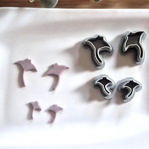 Peut inclure: Ensemble de six découpoirs et moules en argile en forme de raies manta. Les découpoirs sont gris avec des intérieurs noirs, tandis que les pièces d'argile sont de couleur violet clair. Les articles sont disposés sur une surface blanche.