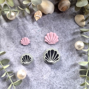 Könnte beinhalten: Eine Sammlung von muschelförmigen Objekten in Rosa und Grau, zusammen mit echten Muscheln und Perlen, angeordnet auf einer grauen Oberfläche. Die rosa Muscheln sind in verschiedenen Größen erhältlich. Die grauen Muscheln haben ein schwarzes Innenleben. Grünzeug umgibt die Objekte.