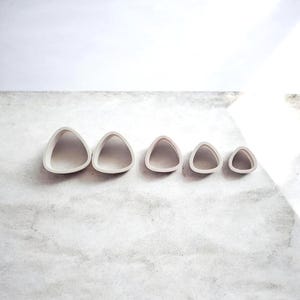 Könnte beinhalten: Ein Satz von fünf hellgrauen, dreieckigen Ausstechern in verschiedenen Größen. Die Ausstecher sind in einer Reihe angeordnet, wobei die Größe von links nach rechts abnimmt. Sie bestehen aus einem glatten, matten Material und sind vor einem hellgrauen Hintergrund platziert.