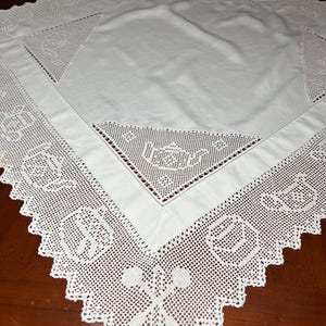 May include: A square white tablecloth featuring a central fabric panel surrounded by an intricate crocheted border. The lace border displays repeating patterns of teapots, cups, and crossed spoons, finished with a scalloped edge.