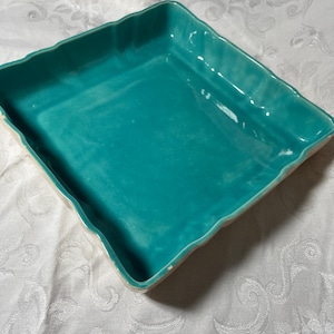 May include: A square ceramic baking dish with a glossy teal interior and a scalloped, wavy rim. The exterior of the dish features a neutral, light-colored finish. The vessel has a deep basin and a distinct, undulating edge design throughout.
