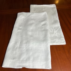 May include: Two rectangular white cloth napkins featuring a subtle, woven damask pattern. The fabric displays a repeating tonal design with intricate floral motifs and a distinct geometric Greek key border detail along the lower edge of the cloth.