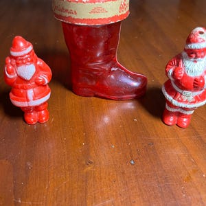 May include: Two small Santa Claus figurines in red and white, flanking a translucent red glass boot container. The boot features a tan paper band around the top rim with the printed text Christmas and additional illegible small lettering.