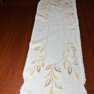 Puede incluir: Un camino de mesa rectangular blanco con patrones de hojas bordadas en tonos amarillo, dorado y marrón. El camino tiene un borde festoneado acabado con encaje de ganchillo blanco y pequeños detalles bordados circulares en las esquinas.
