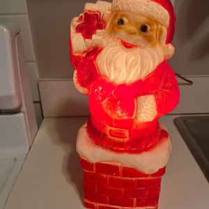 May include: A vintage Christmas decoration of Santa Claus in a red suit and hat, carrying a sack of gifts. The figure is lit with a warm red light and stands on a red brick chimney with a white, snow-like trim.